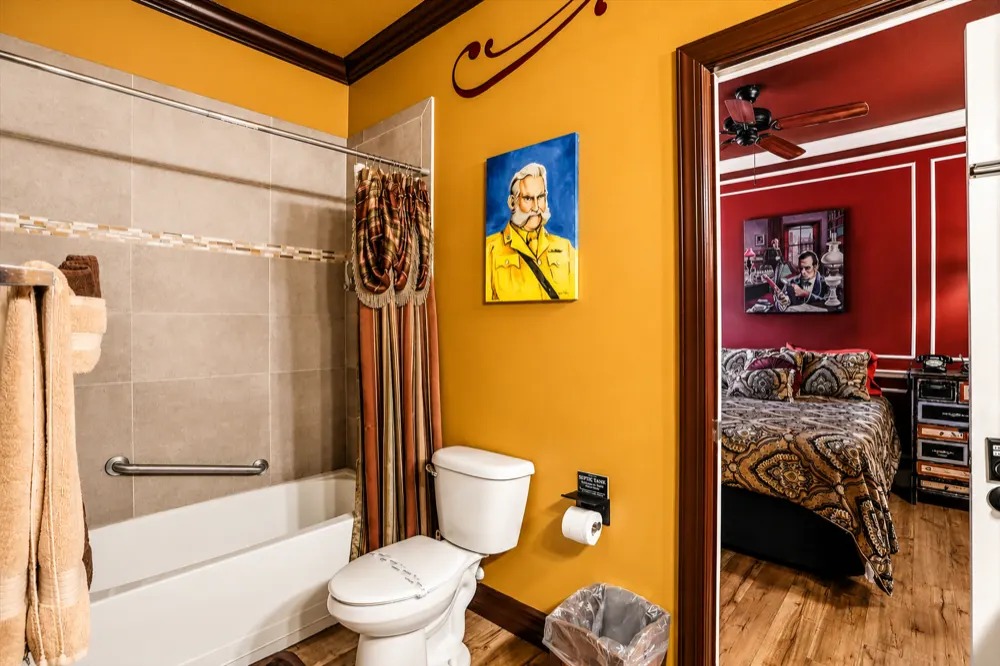 The Clueless bath from the tub side: a Colonel Mustard portrait on the mustard wall, the tiled tub and shower, the Get A Clue room through the door