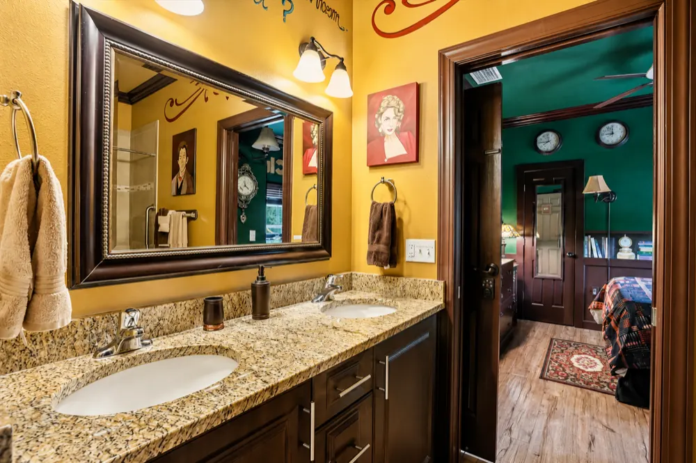 The Clueless bath: mustard walls, a dark double vanity with granite, a framed mirror, a portrait of Miss Scarlet, the green Clue Me In room through the door