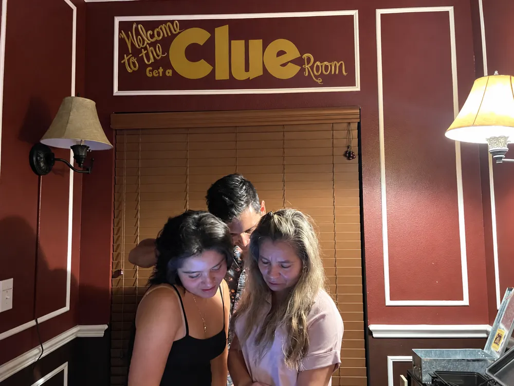 Three guests reading a clue together under the Welcome to the Get A Clue Room sign, the blinds drawn and the lamps lit