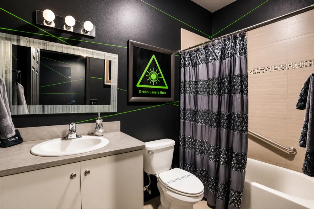 The Laser Games bath with the lights on: black walls with green beam lines, a Green Lasers Rule sign, a sequined shower curtain over the tub, a granite vanity