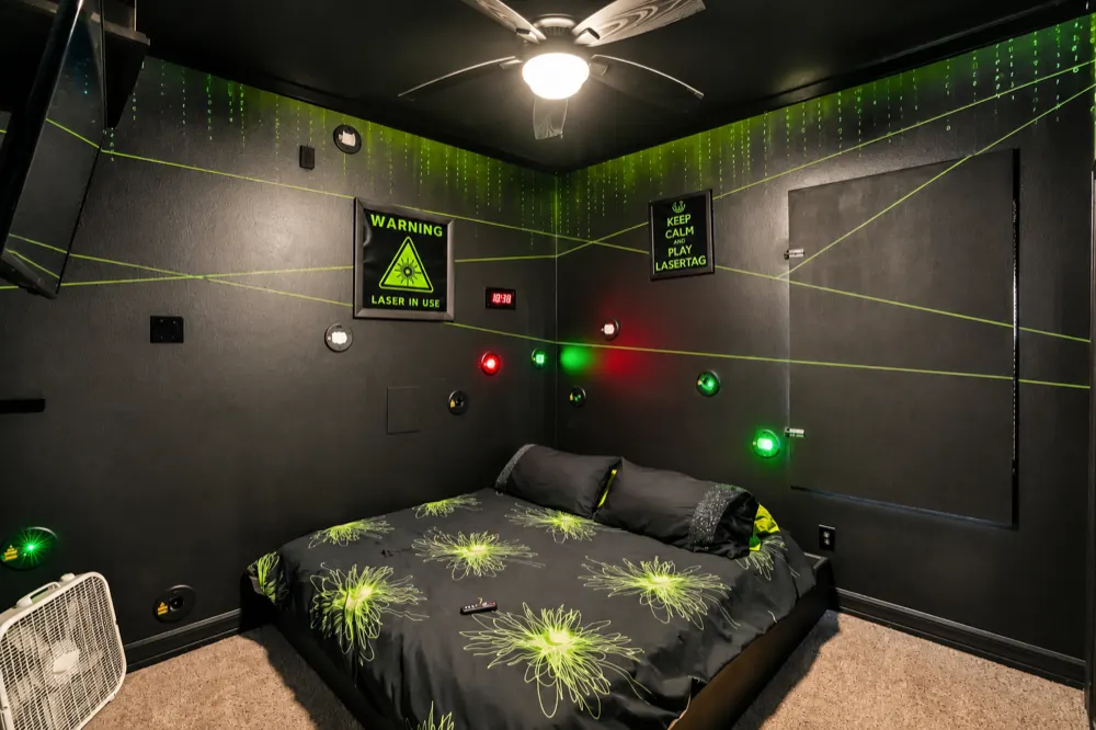 The Laser Games room with the lights on: a king bed with green-burst bedding, the WARNING LASER IN USE sign, the red timer and the Keep Calm And Play Lasertag sign