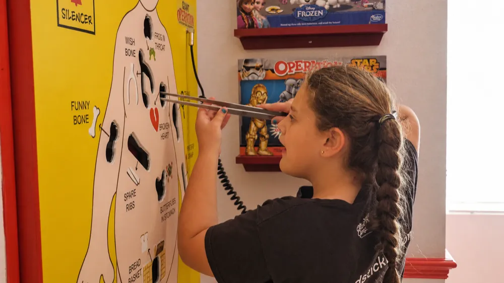 A teenager leaning in with the tweezers on the giant Operation board, the small Operation editions on the shelf beside it