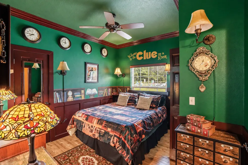Clue Me In bedroom at Great Escape Lakeside: green walls, a row of wall clocks, a painted bookshelf, a stained-glass lamp and the queen bed