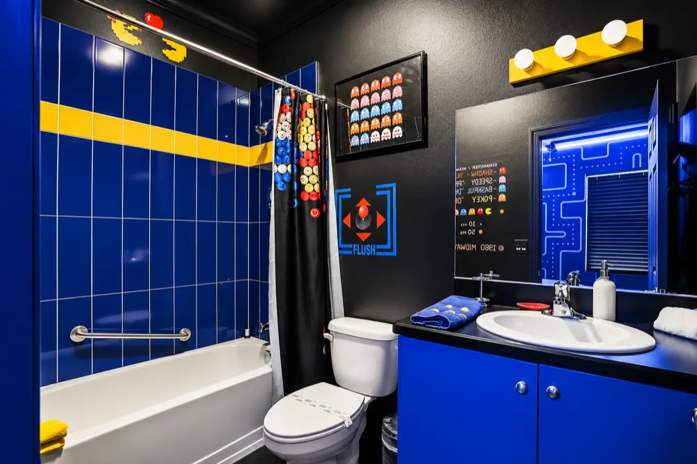 The 80s arcade bathroom: blue tile with a yellow pellet stripe, a Space Invaders shower curtain, a FLUSH joystick decal and a Pac-Man maze above the mirror