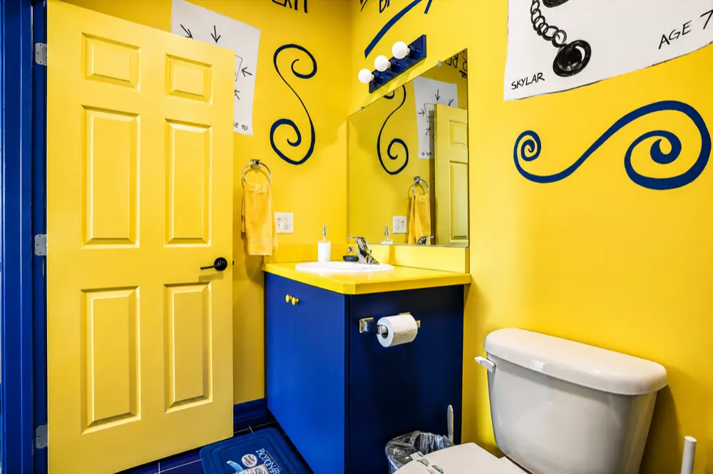 The Pictionary bath: yellow walls with navy marker swirls, a yellow door, a navy vanity with a yellow top, guest drawings taped above the mirror